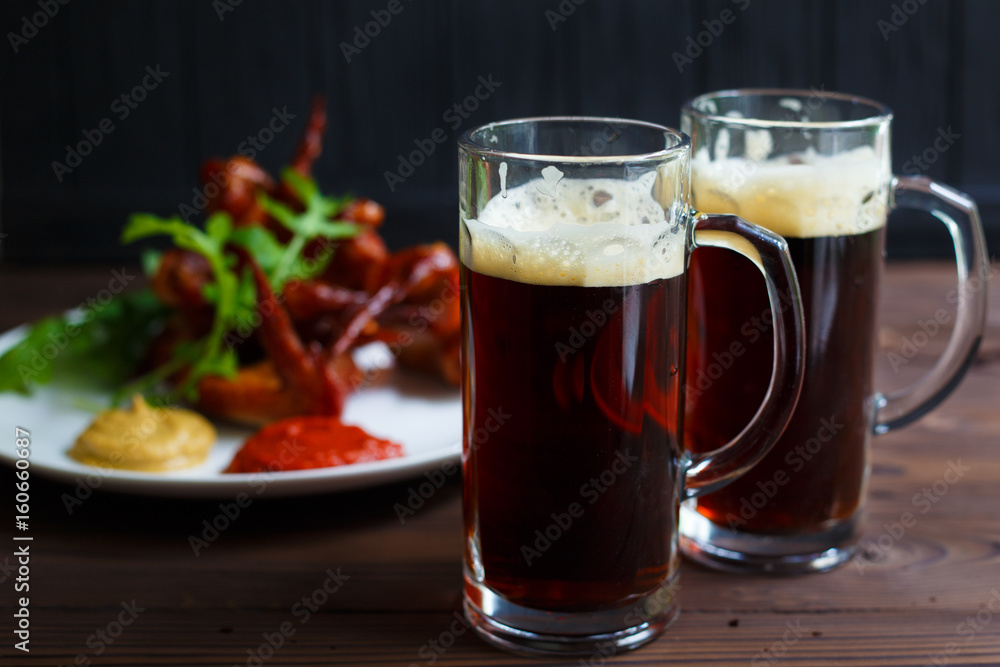 Two mugs of dark beer with delicious hot crusty grilled chicken wings with mustard and spicy tomato sauce Two mugs of dark beer with delicious hot crusty grilled chicken wings with mustard and spicy tomato sauce
