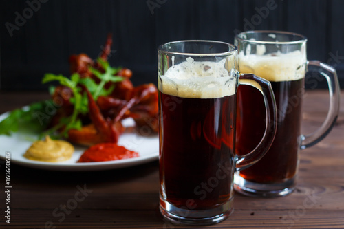 Two mugs of dark beer with delicious hot crusty grilled chicken wings with mustard and spicy tomato sauce