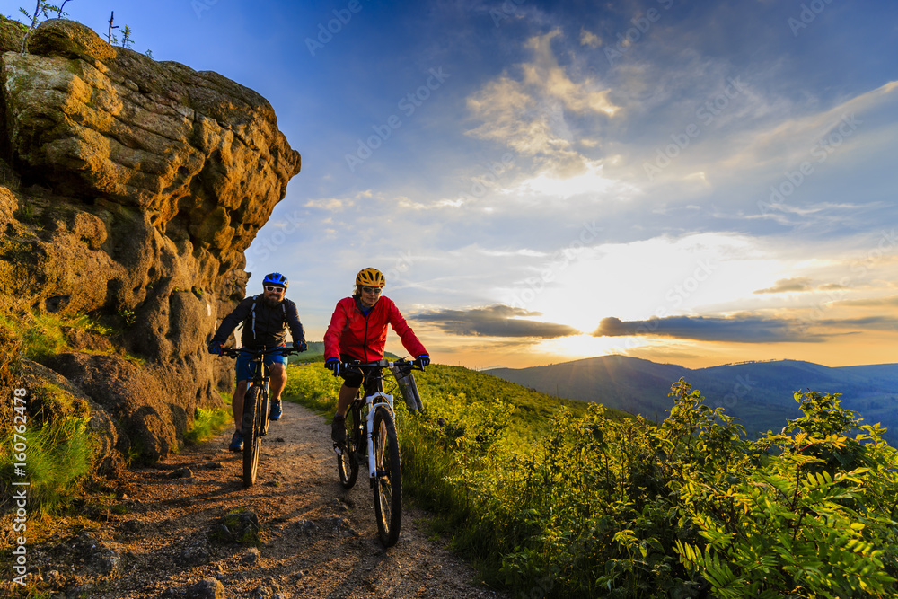 Fototapeta premium Mountain biking women and man riding on bikes at sunset mountains forest landscape. Couple cycling MTB enduro flow trail track. Outdoor sport activity.