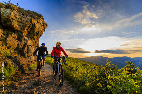 Fototapeta Naklejka Na Ścianę i Meble -  Mountain biking women and man riding on bikes at sunset mountains forest landscape. Couple cycling MTB enduro flow trail track. Outdoor sport activity.