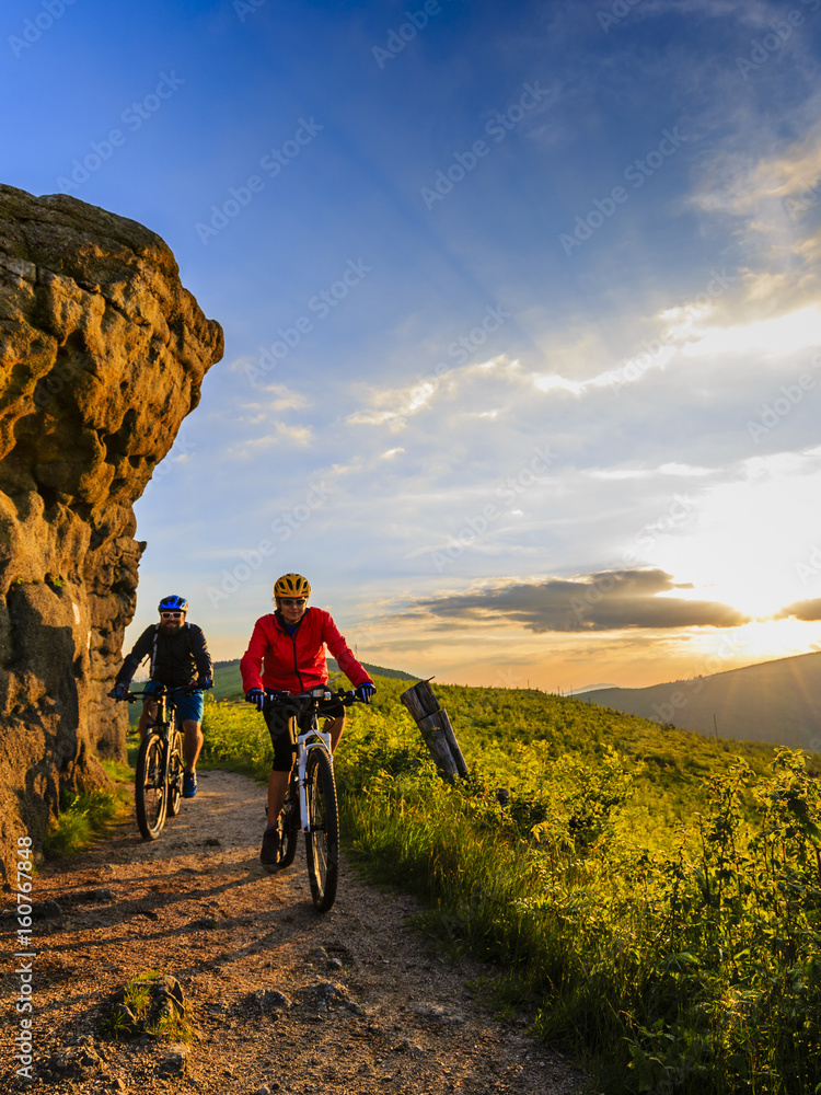 Fototapeta premium Mountain biking women and man riding on bikes at sunset mountains forest landscape. Couple cycling MTB enduro flow trail track. Outdoor sport activity.