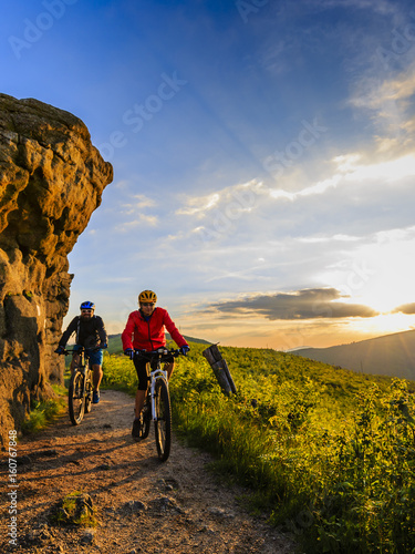 Mountain biking women and man riding on bikes at sunset mountains forest landscape. Couple cycling MTB enduro flow trail track. Outdoor sport activity.