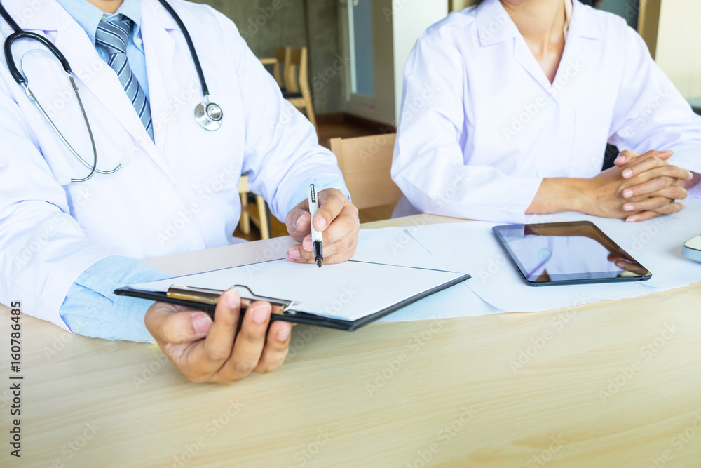 Two doctors discussing patient notes in an office pointing to a clipboard with paperwork as they make a diagnosis or decide on treatment Two doctors discussing patient notes in an office pointing to a clipboard with paperwork as they make a diagnosis or decide on treatment