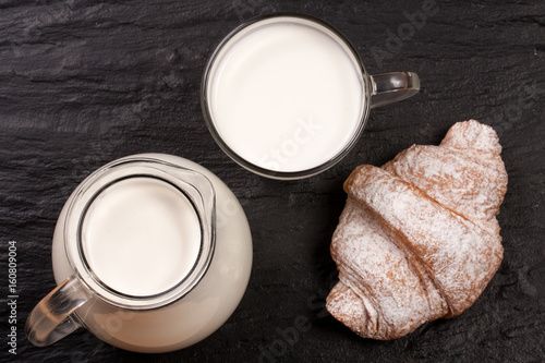 Jug and a glass of milk with a croissant on a black stone background. Top view