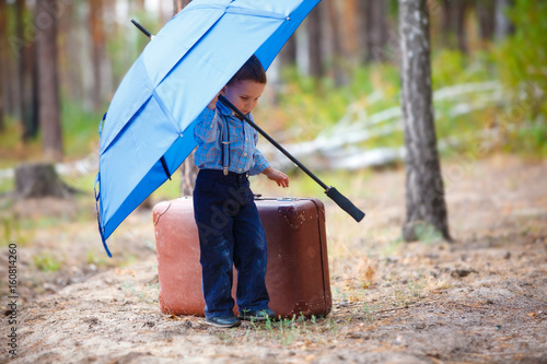 The boy stands in the middle of a pine forest near a vintage leather suitcase and holds a huge blue umbrella over himself