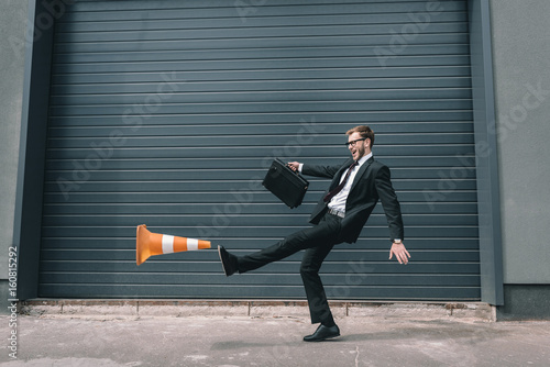 Foto Excited businessman in eyeglasses holding briefcase and kicking traffic cone