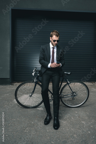 Stylish young businessman in sunglasses sitting on bicycle and using smartphone