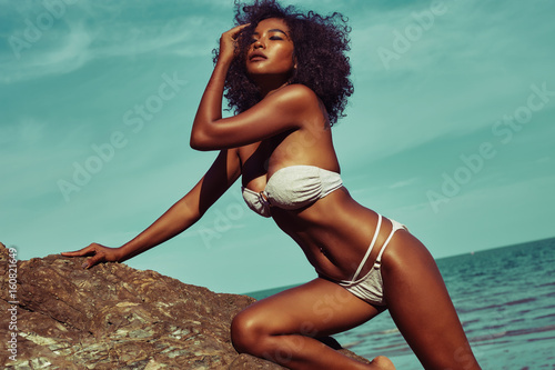 Exotic model with tanned skin and curle hair in bikini posing at the rock beach photo