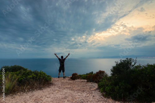 Young man with arms up on the edge of a cliff enjoying a cloudy sunset seascape