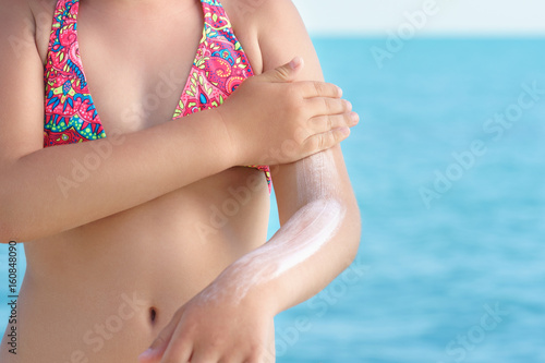 young girl applyng sun protector cream at her shoulder on the beach close to tropical turquoise sea under blue sky