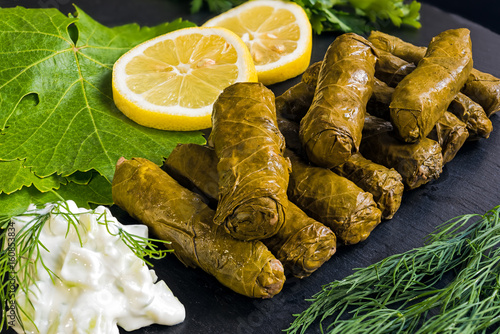 Delicious stuffed grape leaves (the traditional dolma of the mediterranean cuisine) on black dish with leaves, lemon slices, dill and tzatziki sauce.