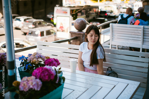 Cute asian young woman in summer cafe outdoors. girl In white T-shirt, with long hair in simple light cozy interior of restaurant Urban style, drinking coffee drink, sitting at table with flowers