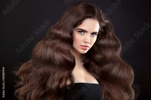 Beauty hair. Brunette girl with long shiny wavy hair. Beautiful girl model portrait with blowing curly hairstyle and clear skin makeup isolated on studio dark background.