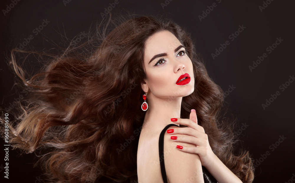 Fototapeta premium Beauty hair. Red manicure. Brunette girl with makeup and long shiny wavy hair. Fashion earring jewelry. Beautiful model portrait with curly hairstyle, manicured nails over dark background.