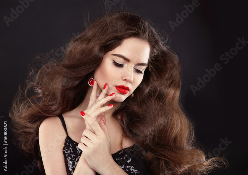 Manicure. Red lips makeup. Beautiful Brunette girl with long blowing shiny wavy hair. Fashion earring jewelry. Elegant model with manicured nails over black background.