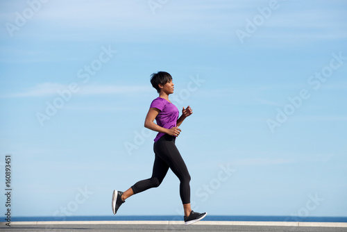 Full body side fit young african woman running outdoors against blue sky