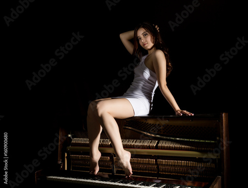 beautiful young woman in a white dress with an open back sitting on a old piano on a dark background photo