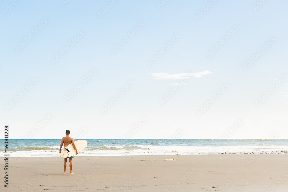 Handsome man in blue swimming shorts and cap stand with long surf surfing board wait on surf spot at sea ocean beach. White blank surfboard. Concept of power, freedom, new modern life, generation Y.