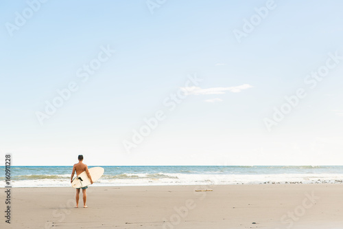Handsome man in blue swimming shorts and cap stand with long surf surfing board wait on surf spot at sea ocean beach. White blank surfboard. Concept of power, freedom, new modern life, generation Y.
