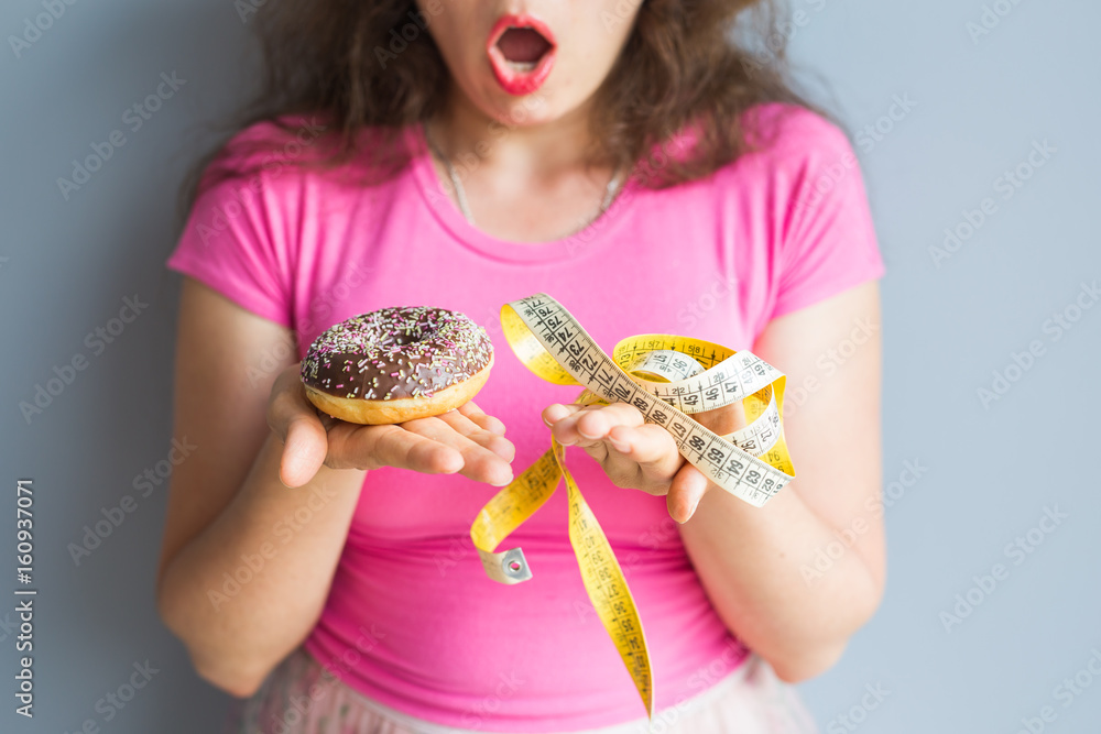 Close-up of confused young woman holding a donut and a measuring tape. The concept of healthy eating. Diet. Close-up of confused young woman holding a donut and a measuring tape. The concept of healthy eating. Diet.