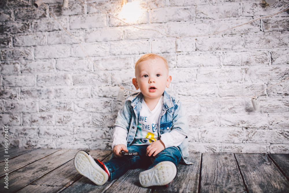 A stylish child, a boy, one year old, sit on a wooden floor and on a brick wall background. He is dressed in a junky jacket, junks and a white T-shirt. Blonde A stylish child, a boy, one year old, sit on a wooden floor and on a brick wall background. He is dressed in a junky jacket, junks and a white T-shirt. Blonde