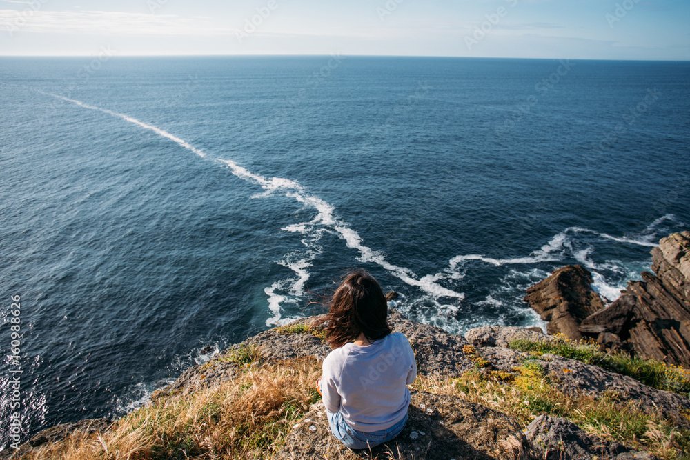 Young woman with dark hair sits on edge of cliff, dreamingly overlooks ocean waves and horizon, makes photo of herself and nature