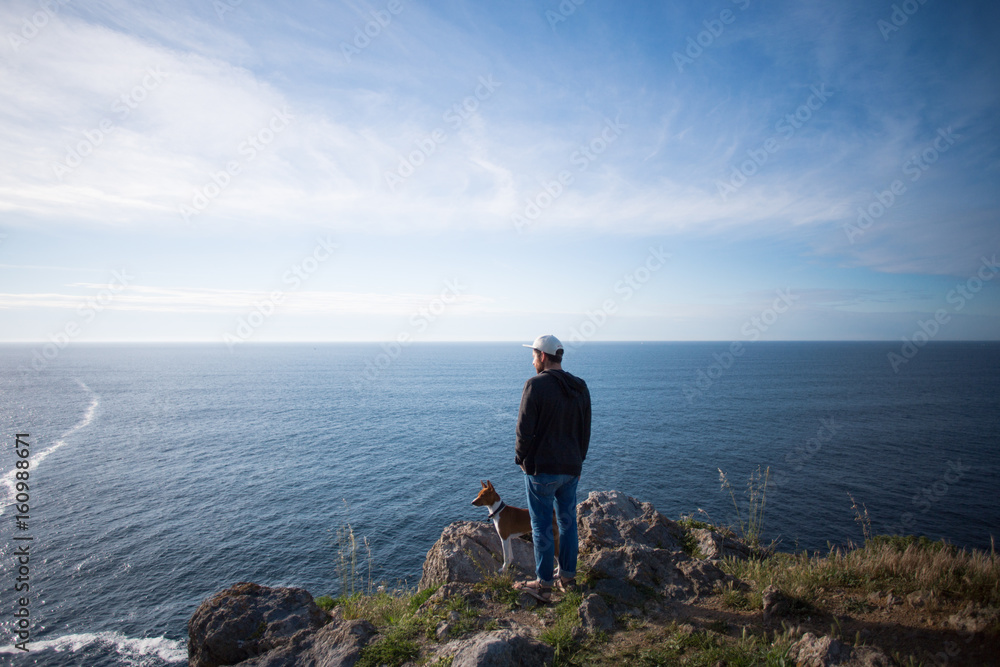 Tall young man in urban casual clothing, stands on edge of mountain and overlooks ocean views on sunset together with best friend, basenji little puppy
