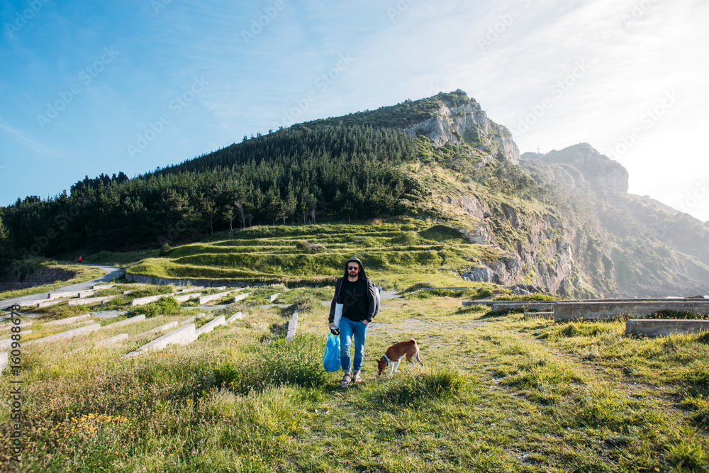 Urban nomad adventurer with backpack walks next to seaside cliffs and mountains in amazing nature scenery with best friend dog of basenji pure breed