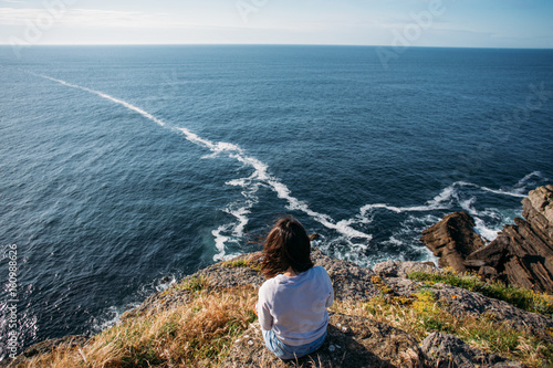 Young woman with dark hair sits on edge of cliff, dreamingly overlooks ocean waves and horizon, makes photo of herself and nature