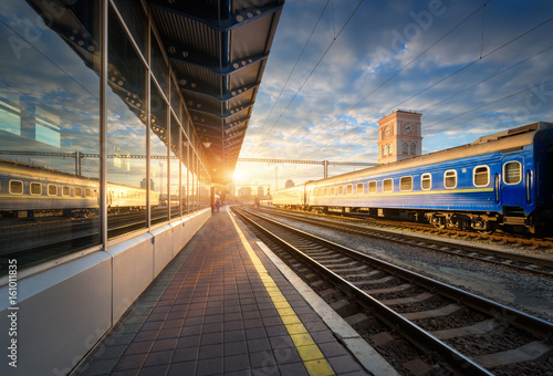 Beautiful blue passenger train at the railway station at sunset. Industrial view with modern train, railroad, railway platform, buildings and blue sky with clouds in the evening. Transportation