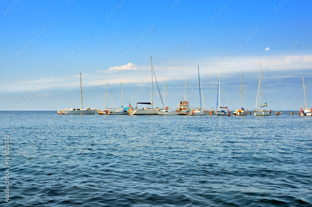 Yachts and sailing boats are on the dock. The small naval vessels are in the harbor on a background of blue sky.