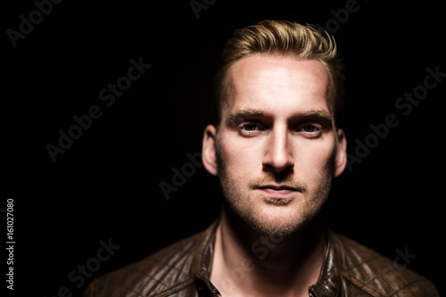 Handsome blonde man standing against a black background with a simple light above his head, wearing a brown leather jacket and white t-shirt.