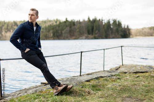 Laid back blonde man in a blue jacket and jeans, leaning back against a railing outdoors on a sunny spring day with the lake behind him.