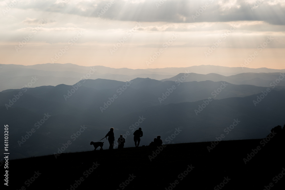 Some people and a dog on top of a mountain, with other mountains and hills in the background, and sunrays coming out of the clouds