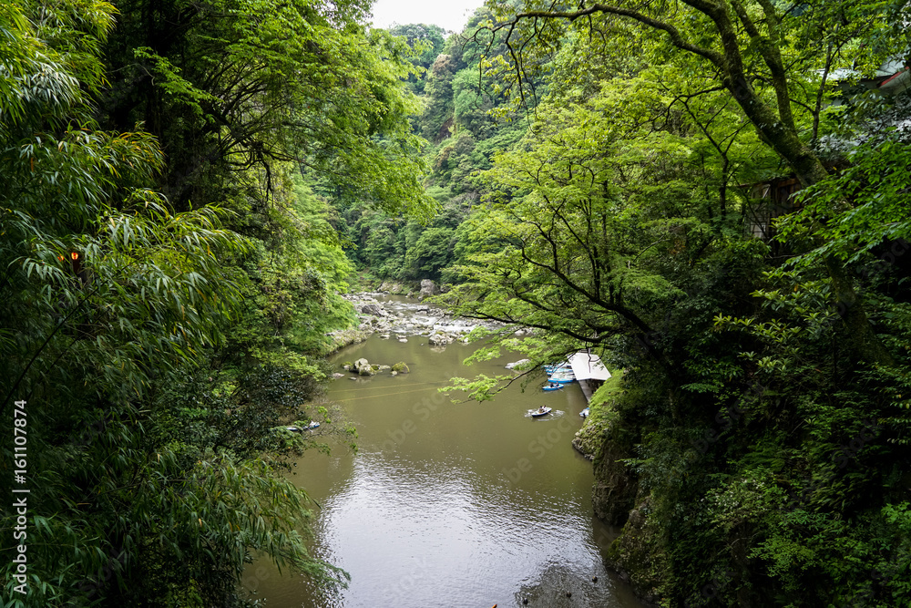 View of Takachiho gorge from above seeing river, boats at pier and rocky shore through lush green trees and mountain background View of Takachiho gorge from above seeing river, boats at pier and rocky shore through lush green trees and mountain background