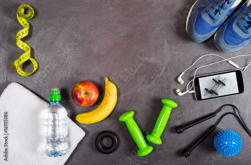 Dumbbell, apple, water bottle, towel, smartphone and headphones, measuring tape, sneakers on grey background. Top view fitness equipment background