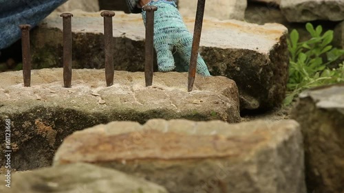 A stonecutter or stonemason is hammering several chisels on the cutting line of a stone. He is cutting the stone in a traditional way, manually. Closeup.