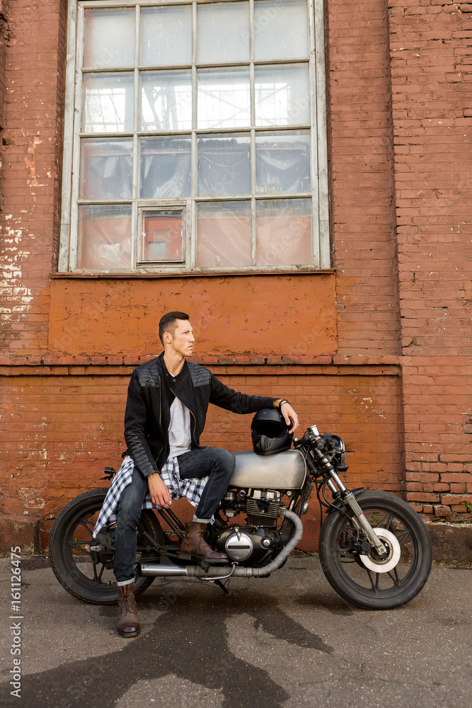 Handsome biker guy in black leather jacket sit on classic style cafe racer motorcycle and hold helmet on gas tank. Bike custom made in vintage garage. Brutal fun urban lifestyle. Outdoor portrait.