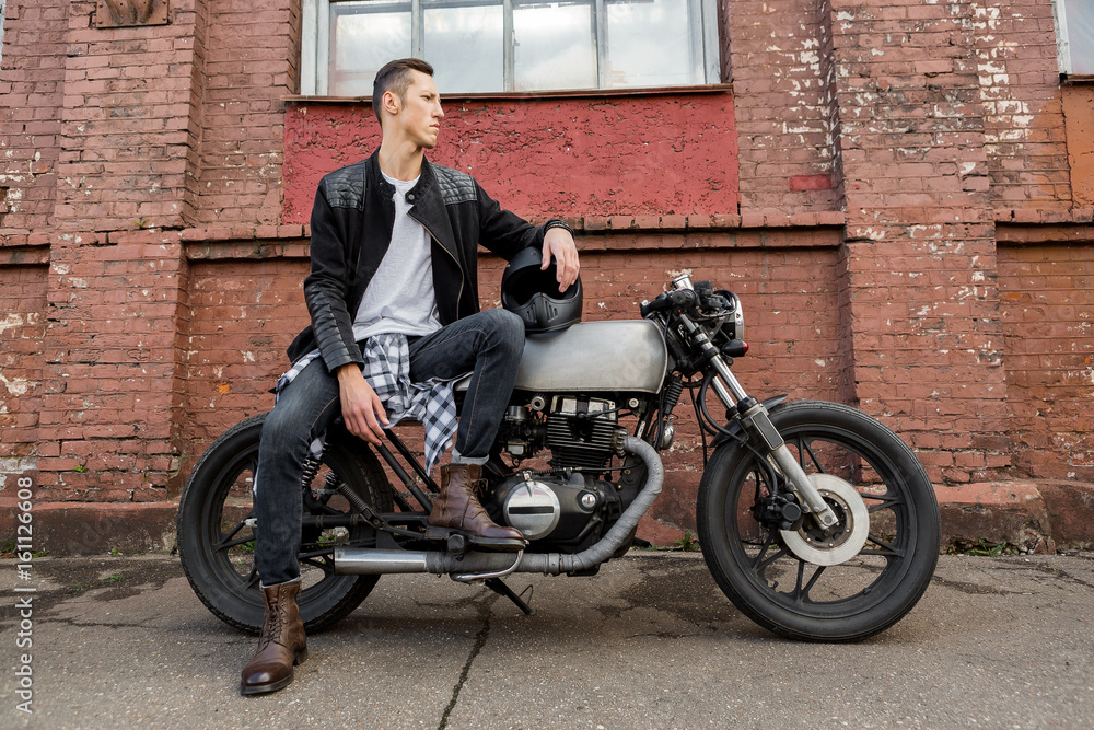Handsome rider biker man in black leather jacket, jeans, boots and helmet sit on classic style cafe racer motorcycle. Bike custom made in vintage garage. Brutal fun urban lifestyle. Outdoor portrait.