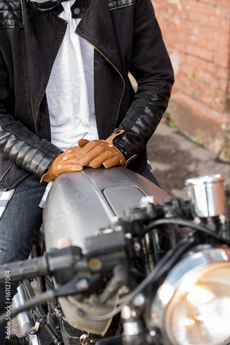 Close up of a hipster biker man hands in leather glove on a raw metal gas tank of classic style cafe racer motorcycle. Bike custom made in vintage garage. Brutal fun urban lifestyle. Outdoor portrait.