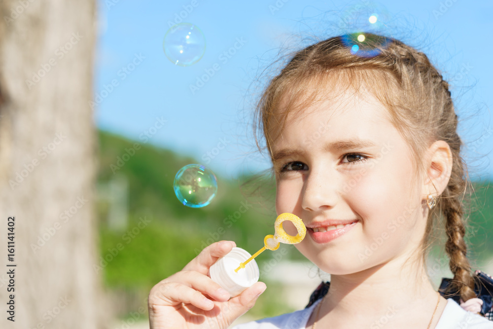 young pretty european kid girl with cute smile is blowing bubbles in outdoor under blue sky at sunny day