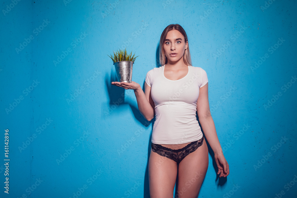 Foto de Sexy blonde woman in a white T-shirt and panties holds a pot with a  flower in her hands. do Stock