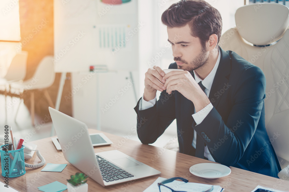 Young man is concentrated on reading his email while drinking coffee in office. He is in formal wear, the office is modern and bright Young man is concentrated on reading his email while drinking coffee in office. He is in formal wear, the office is modern and bright