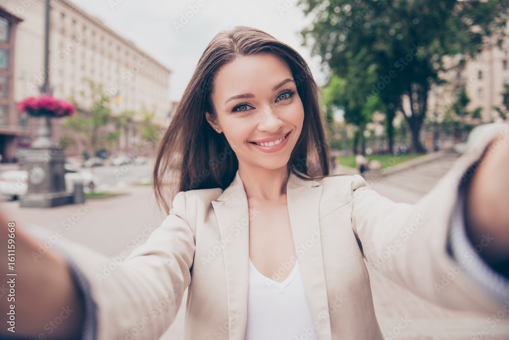 Carefree and happy, sunny spring mood. Charming young lady is making selfie on a camera. She is wearing formal wear, smiling, while on a walk in town outdoors