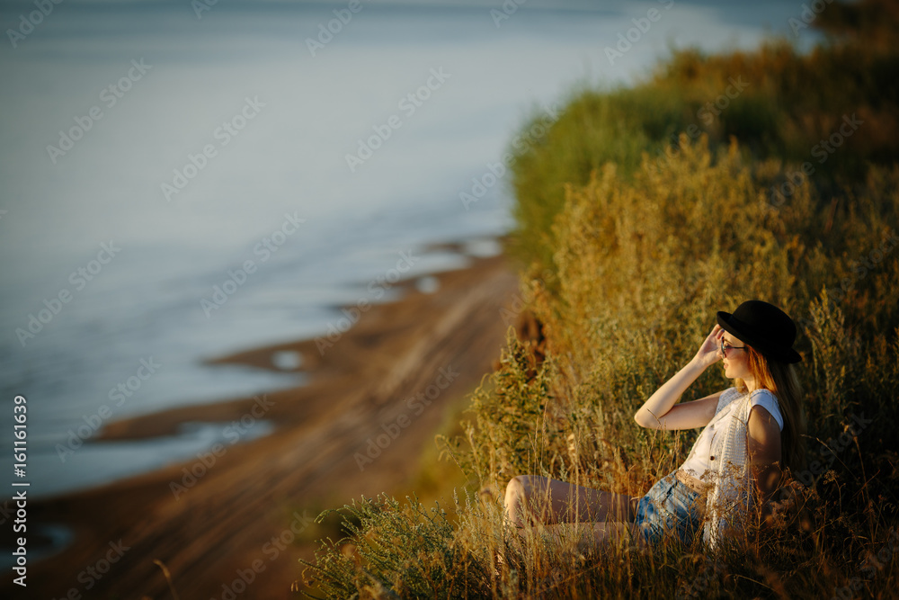 Young pretty woman wearing hat and sunglasses dressed in boho style clothes spending time on the nature on the bluff near river during beautiful sunset