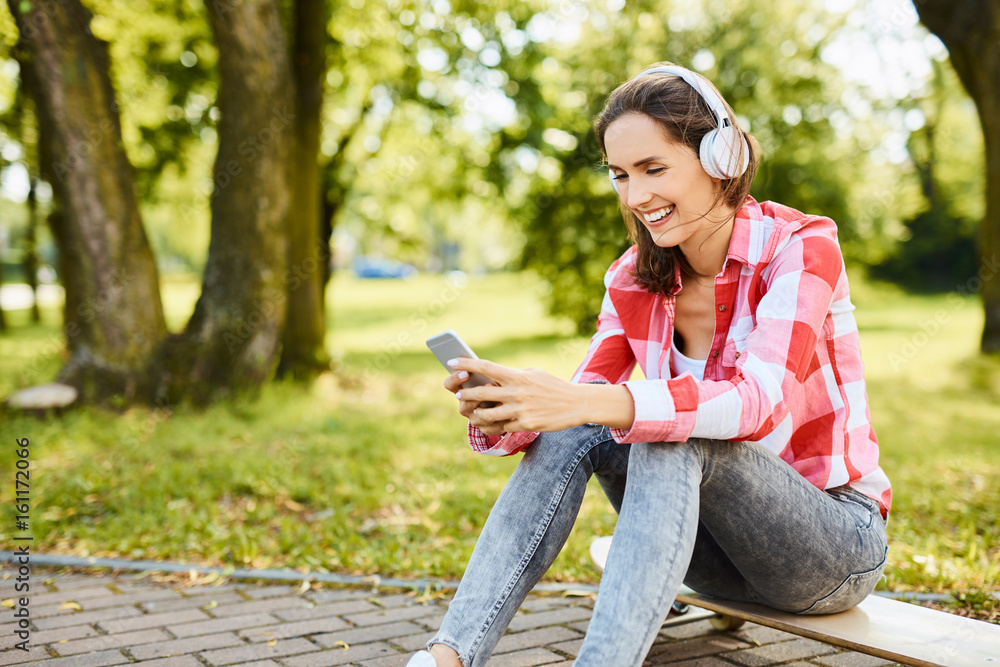 Cheerful young woman siting on longboard, streaming musing on her smart phone Cheerful young woman siting on longboard, streaming musing on her smart phone