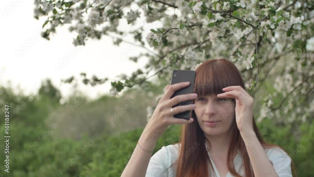 Girl makes selfie in the garden. An attractive red-haired woman smiles making selfi using a mobile phone in a cherry orchard. The concept of using gadgets for a healthy lifestyle. Girl makes selfie in the garden. An attractive red-haired woman smiles making selfi using a mobile phone in a cherry orchard. The concept of using gadgets for a healthy lifestyle.