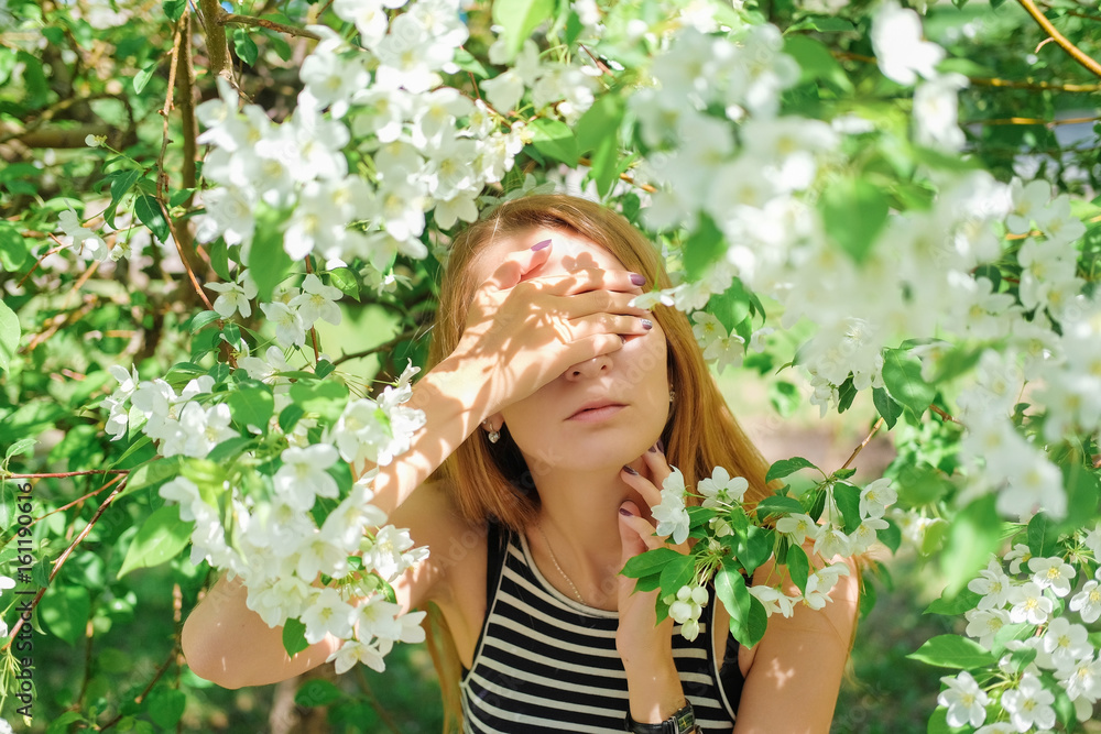 Portrait of a beautiful sensual girl in a dress striped in the background of the apple tree. Summer and spring trend in fashion. A woman covers her eyes with her hands. Portrait of a beautiful sensual girl in a dress striped in the background of the apple tree. Summer and spring trend in fashion. A woman covers her eyes with her hands.