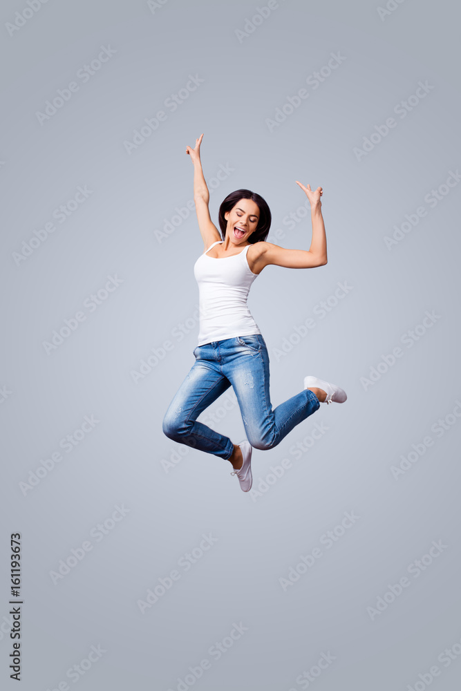 Freedom, carefree, fun, summer mood. Excited young brunette is jumping and showing peace signs. She is in jeans and casual white singlet