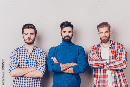 Ethnicity, multicultural diversity. Three serious harsh men are standing isolated on pure background with crossed hands, dressed in casual clothes